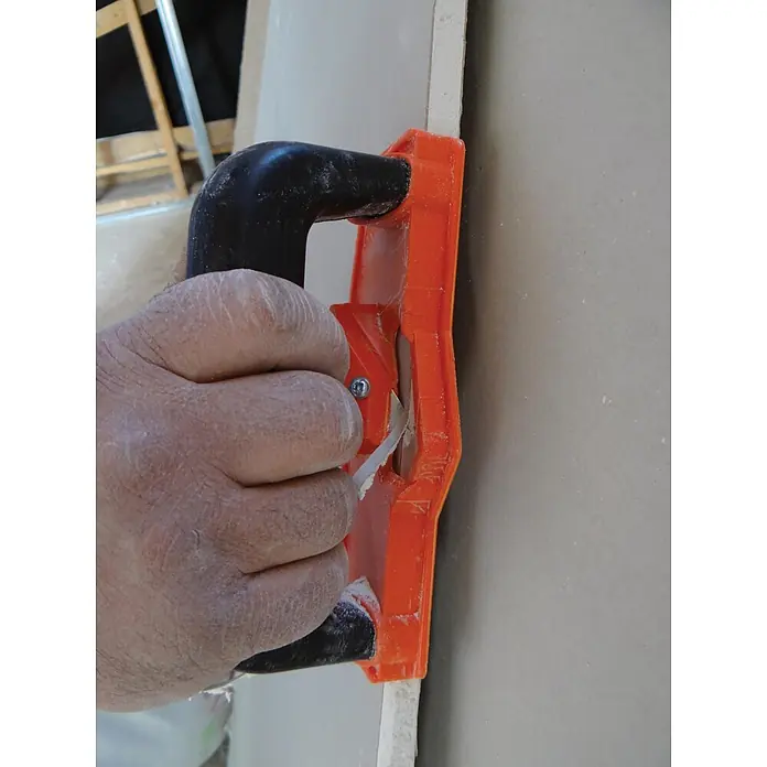 Mondelin Kantenhobel - Geeignet für: Gipskartonplatten - Hand trimmt mit einem orange-schwarzen Gipskartonkantenfräser die Kante einer Gipskartonplatte.