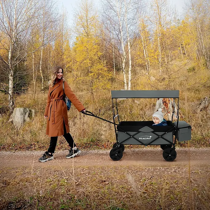 Hauki Bollerwagen Handwagen Transportwagen faltbar klappbar mit Bremse Dach und Tasche - L x B x H: 170 x 53 x 104 cm - Frau zieht grauen HAUKI Bollerwagen mit Verdeck, in dem ein Kind sitzt, auf einem Weg.