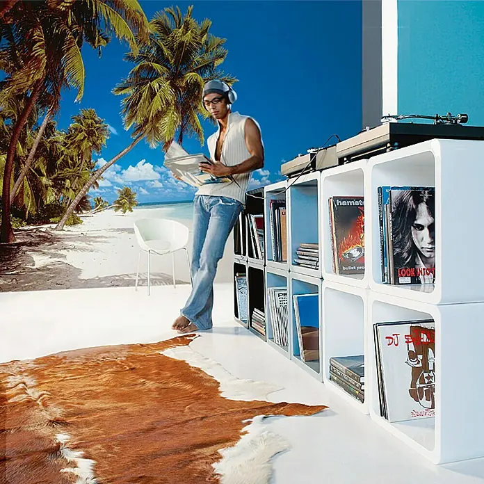 Komar Fototapete Malediven - 8 -tlg., B x H: 388 x 270 cm, Papier - Mann liest Zeitung neben weißem Schallplattenregal, darauf ein Plattenspieler. Im Hintergrund eine Strandtapete und ein Kuhfellteppich.
