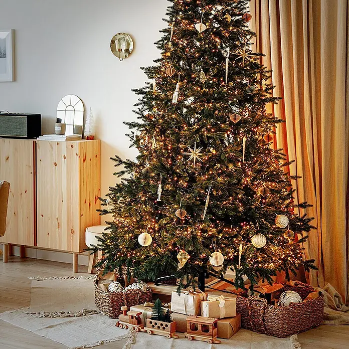 ECD Germany LED-Clusterlichterkette 1512 LEDs 11m für Innen & Außen IP44 Warmweiß - Außen, 1.512 Stk., Netzbetrieben - Ein hoher, hell erleuchteter Weihnachtsbaum, geschmückt mit warmen Lichtern und goldenen Ornamenten, umgeben von Geschenken und Weidenkörben.
