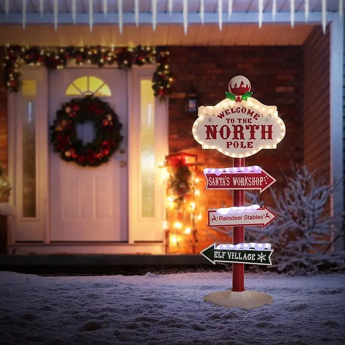 ECD Germany LED-Weihnachtsdekofigur LED Wegweiser Weihnachtsbeleuchtung Nordpol Schild Deko - Weiß, Rot, Grün, Netzbetrieben, LED - Ein beleuchteter rot-weißer Weihnachts-Wegweiser in einem verschneiten Garten, der Richtungen wie 'Nordpol' und 'Santas Werkstatt' anzeigt, mit Lichtern geschmückt.