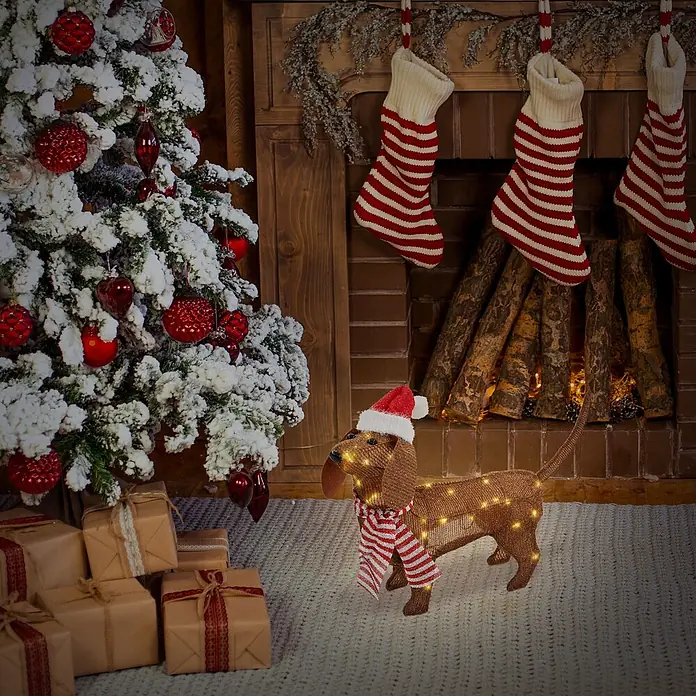 ECD Germany LED-Weihnachtsdekofigur LED Weihnachten Hund Dackel Gartenfigur Hundefigur Batteriebetriebene - Braun, Akkubetrieben, LED - Eine braune, beleuchtete Dackel-Weihnachtsdekoration mit Weihnachtsmütze und gestreiftem Schal, auf einem Teppich in einem festlichen Raum.