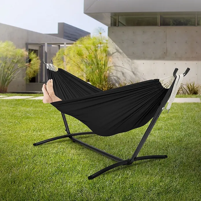 ML-Design Hängematte XXL Hängesessel Schwarz mit Tragetasche und Ständer für Garten/Balkon - Stahl, Schwarz, Passend für: Erholung, Lesen, Entspannen - Schwarze Stoffhängematte auf schwarzem Metallgestell draußen, mit Füßen in der Hängematte.