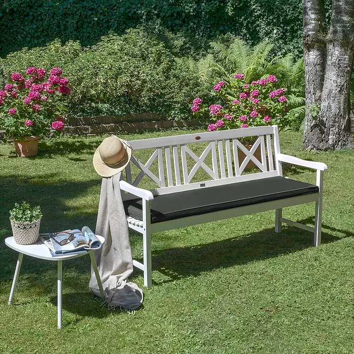 Beautissu Bankauflage Loft BK - Gartenbank, 1 Stk., L x B x H: 100 x 48 x 5 cm, Polyester, Graphitgrau - Weiße Holzgartenbank mit dunkelgrauem Kissen, Hut und Decke, neben einem Beistelltisch in einem sonnigen Garten.