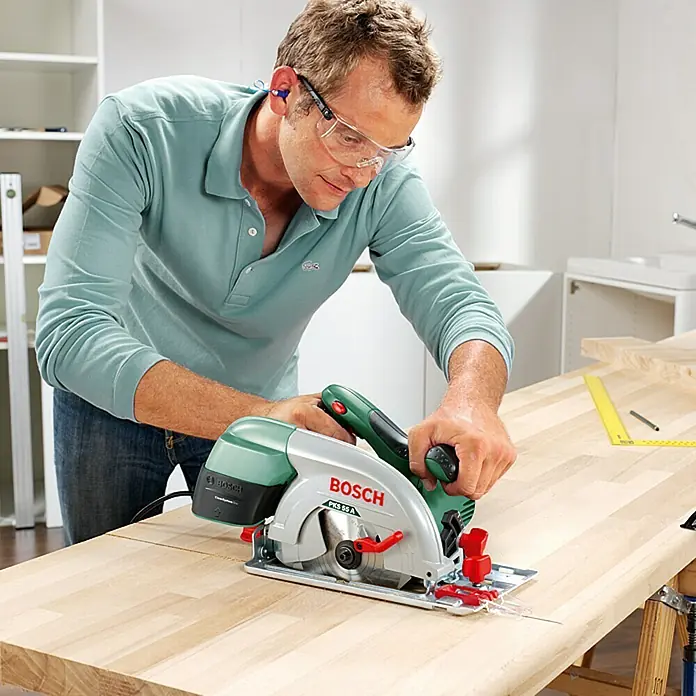 Bosch Handkreissäge PKS 55 A - 1.200 W, 5.600 U/min - Ein Mann benutzt eine Bosch PKS 55 A Handkreissäge auf einer Holzplatte, mit Schutzbrille und Gehörschutz für Sicherheit.