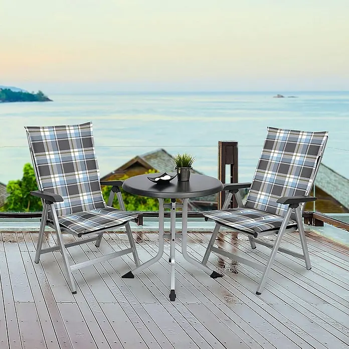 Beautissu Sitzauflage Sunny HL - Hochlehner, 2 Stk., Polyester, Blau - Zwei klappbare Terrassenstühle mit Karomuster und ein runder Tisch auf einer Terrasse mit Meerblick. Graue Metallrahmen.