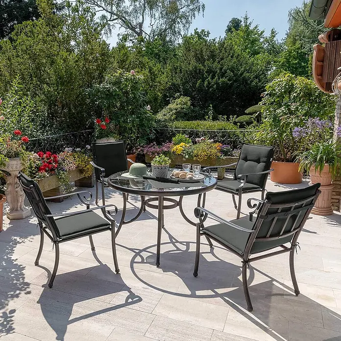Beautissu Sitzauflage Loft Nl - Niederlehner, 8 Stk., L x B x H: 100 x 50 x 6 cm, Polyester, Graphitgrau - Dunkles Metall-Gartenset mit Glastisch und vier Stühlen mit dunkelgrauen Kissen, auf einer hellen Terrasse im üppigen Garten.