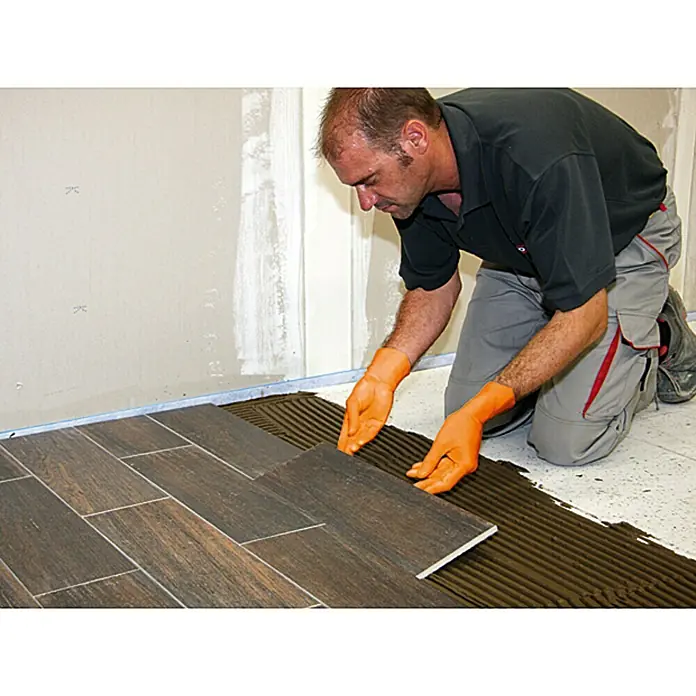 Racofix Flexkleber Super-Flexkleber - 15 kg - Ein Handwerker verlegt sorgfältig eine dunkle Fliese auf einem Boden mit Kleber. Die orangefarbenen Handschuhe schützen seine Hände bei der Arbeit.