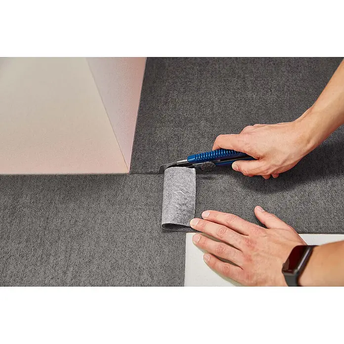 Teppichfliesen 40x40 cm selbstklebend Teppichboden Nadelvlies Grau -  - Hand schneidet mit blauem Teppichmesser dunkles graues Filzunterlagenmaterial. Ein Streifen wird dabei entfernt.