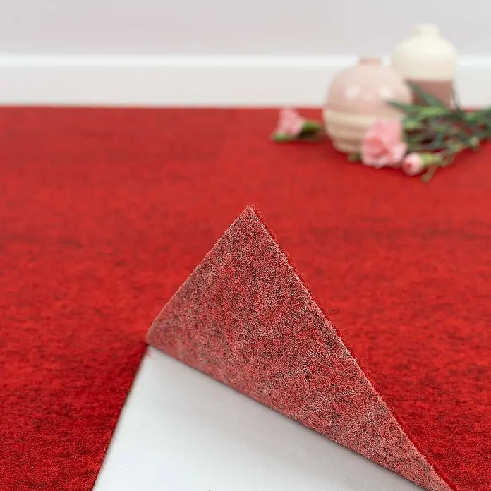 Bricoflor Teppichfliese Nadelvlies Nadelfilz, Rot, 40x40cm -  - Roter Filzteppich wird abgezogen, um die weiße Kleberückseite zu zeigen. Verschwommene Deko im Hintergrund.
