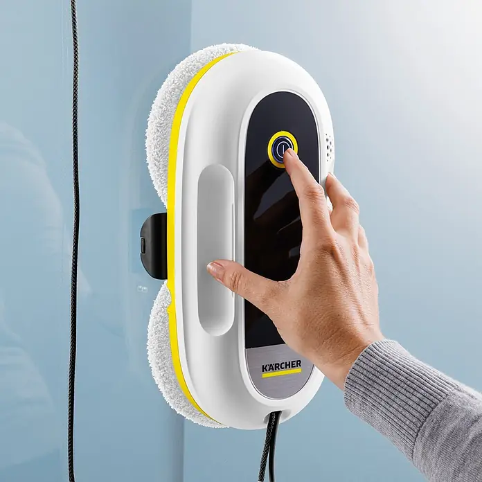 Kärcher Fensterreinigungsroboter RCW 2 - Wassertankvolumen Roboter: 70 ml, Saugkraft: 3.300 Pa - Eine Hand aktiviert einen weiß-gelben Kärcher Fensterputzroboter mit schwarzem Bedienfeld, der an einem Fenster haftet, Sicherungsseil sichtbar.