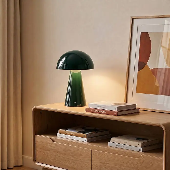 Lindby Tischleuchte Nevijo - grün, grün - Eine dunkelgrüne pilzförmige Tischlampe mit leuchtendem Fuß steht auf einem Holzschrank und beleuchtet Bücher und ein Bild.