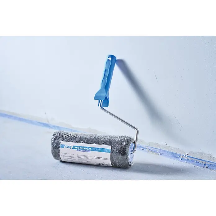 Knauf Spachtelwalze - 240 mm - Knauf Spachtelwalze mit blauem Griff und dunkel, in Plastik verpackter Rolle, lehnt an einer Wand in einer Baustelle.
