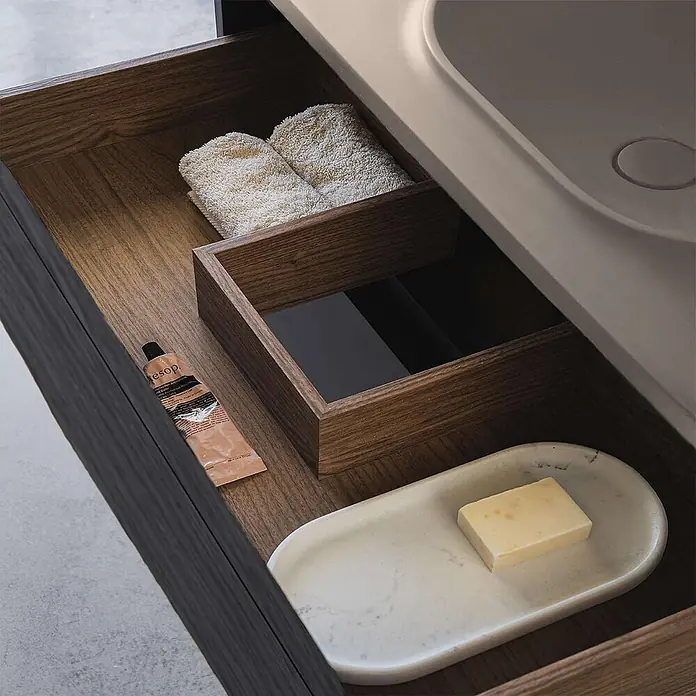 Poalgi Badmöbel-Set BÜL - Gesamtbreite Badmöbel: 80 cm, Nussbaum und Perle, Matt - Geöffnete Holzschublade mit Handtüchern, Aesop-Creme und Marmorseifenschale mit Seife unter einem hellen Waschbecken.