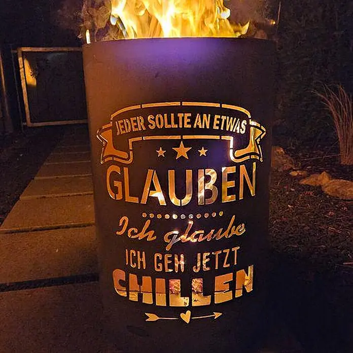 TiKo-Design® Feuertonne Chillen Ø 40 cm – massive Feuerstelle -  - Dunkle Metall-Feuerschale mit leuchtenden deutschen Textausschnitten und aktiven Flammen, draußen in der Nacht.