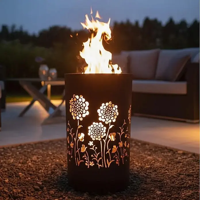 TiKo-Design Holzkohlegrill Feuertonne-Flower Power - grau - Zylindrische dunkle Metall-Feuerstelle mit beleuchteten Blumenausschnitten, hell brennend auf einer Terrasse.