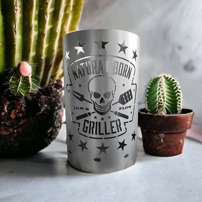 TiKo-Design® Feuertonne Natural Born Griller Ø 40 cm – Grill Motiv -  - Silberfarbener Metallzylinder mit lasergeschnittenem „Natural Born Griller“-Design, Totenkopf, Grillwerkzeugen und Sternen, zwischen zwei Kakteen.