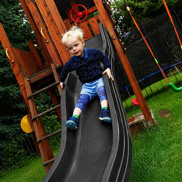 Wellenrutsche Anbaurutsche 3,00 x 0,50 m anthrazit -  - Ein kleines Kind sitzt auf einer dunkelgrauen Spielplatzrutsche in einer Holzspielanlage, bereit zum Rutschen.