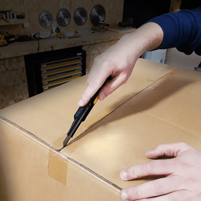 Vito Cúter de seguridad VIX18AA - Anchura de hoja: 18 cm - Mano usando un cúter negro y amarillo para abrir una caja de cartón marrón en un banco de trabajo.