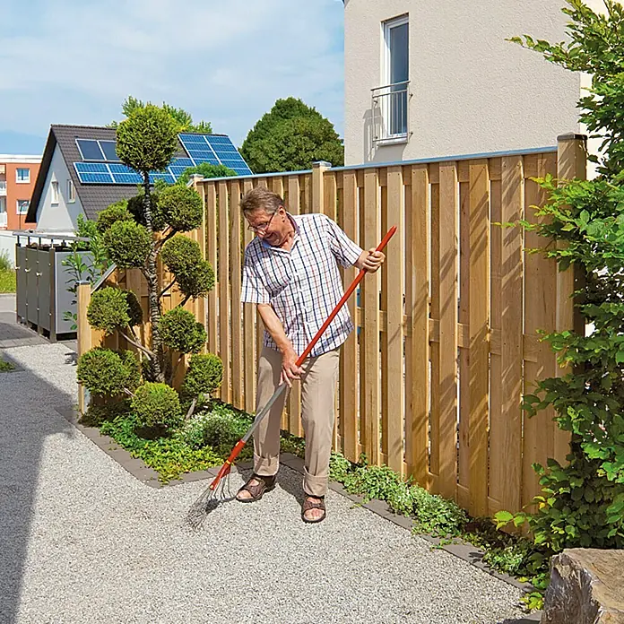 Designaufsatz - Länge: 180 cm, Geeignet für: Sichtschutzzäune von 40 - 45 mm - Ein Mann harkt Kies auf einem Weg neben einem Holzzaun. Der Zaun ist aus hellbraunem Holz und hat eine moderne Optik.
