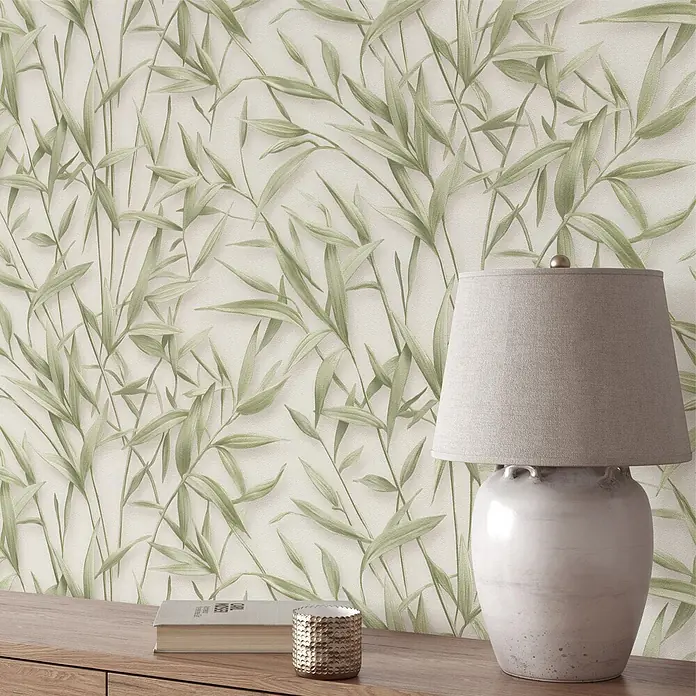 Erismann Papel pintado de vinilo Bambú - Verde/Beige, Floral, 10 x 0,53 m - Papel pintado con patrón de hojas de bambú verdes, mostrado en una pared detrás de una mesa de madera con lámpara.