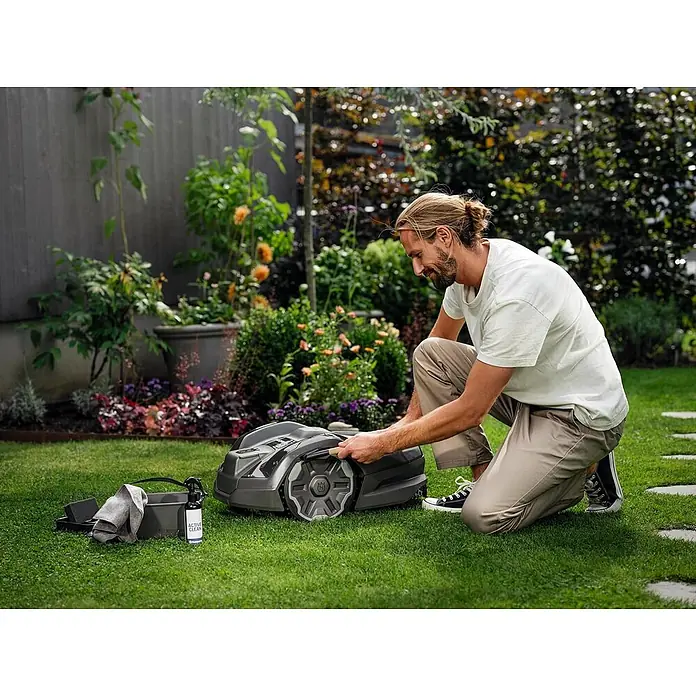 Husqvarna Automower Aspire R6V Mähroboter-Set 600 m² + Cleaning Kit -  - Mann reinigt einen dunkelgrauen Mähroboter und sein Zubehör mit Reinigungsspray und Tuch im Garten.