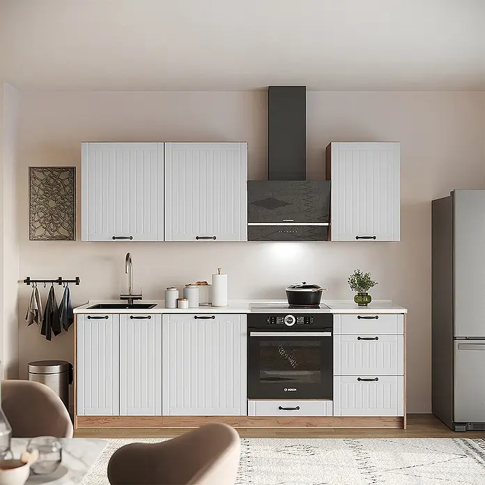 Vicco Küchenzeile - Weiß Landhaus, Ohne Elektrogeräte - Helle Küche mit weißen gerippten Schränken, schwarzen Griffen, integriertem schwarzem Ofen, Dunstabzugshaube und Spüle, dazu ein silberner Kühlschrank.