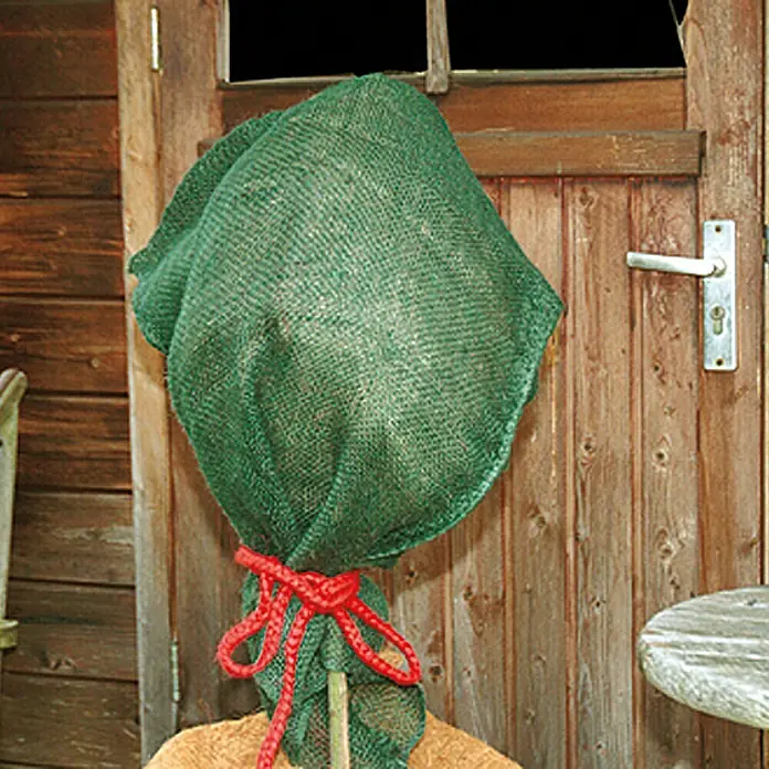 Floraworld Classic Gartensack - Grün, 105 cm - Ein grüner Jutesack schützt eine Pflanze vor einem Holztor, zusammengebunden mit einer roten Schnur.