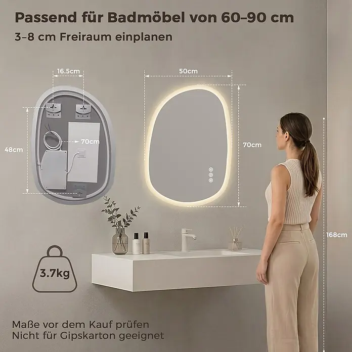 S'AFIELINA Lichtspiegel Mora 700 × 500 mm - 70 x 50 cm - Ein beleuchteter, organisch geformter Badezimmerspiegel (50x70cm) hängt über einem weißen Waschbecken, daneben steht eine Frau zur Größenreferenz.