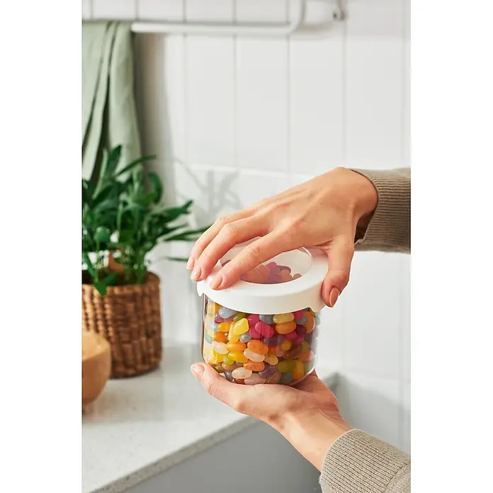 SmartStore Vorratsdose Vision - Ø x H: 11,5 x 10 cm, Kunststoff, Weiß - Hände halten ein klares Glas mit bunten Geleebohnen, gezeigt auf einer Küchenzeile.