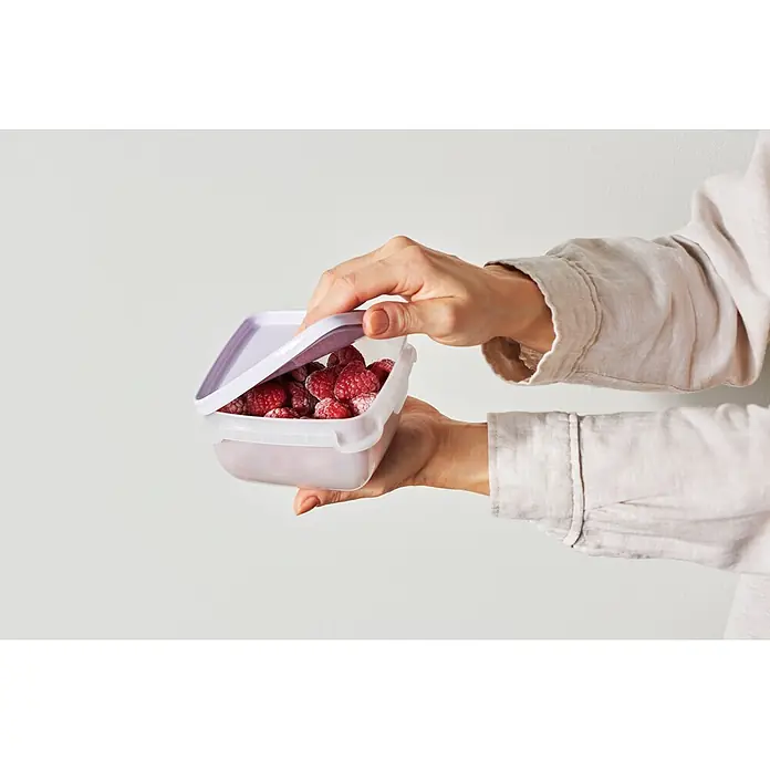 SmartStore Vorratsdosen-Set - L x B x H: 12 x 12 x 6,5 cm, Kunststoff, Weiß/Violett - Hände öffnen eine klare, rechteckige Lebensmittelbox mit lila Deckel, gefüllt mit gefrorenen Himbeeren.