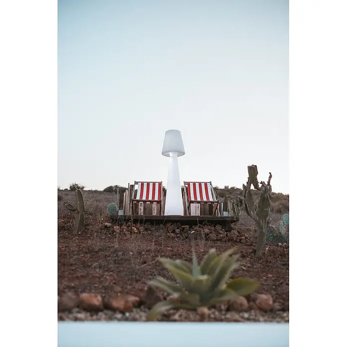 New Garden Lámpara de pie con batería Lola 165 Blanco - Blanco, Altura: 165 cm, RGB - Dos tumbonas a rayas rojas y blancas y una lámpara de exterior blanca alta forman un acogedor rincón en un paisaje seco con cactus.