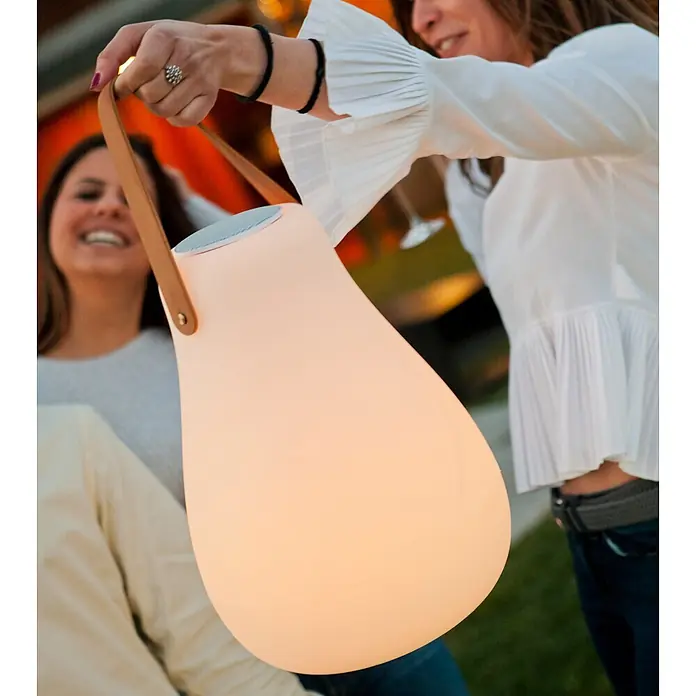 New Garden Lámpara para exterior con altavoz Kurby 38 Play - Blanco, Altavoz Bluetooth, Ø x Al: 27,5 x 37 cm, RGBW - Lámpara/altavoz portátil luminosa en forma de pera con asa de cuero marrón, sostenida por una persona.