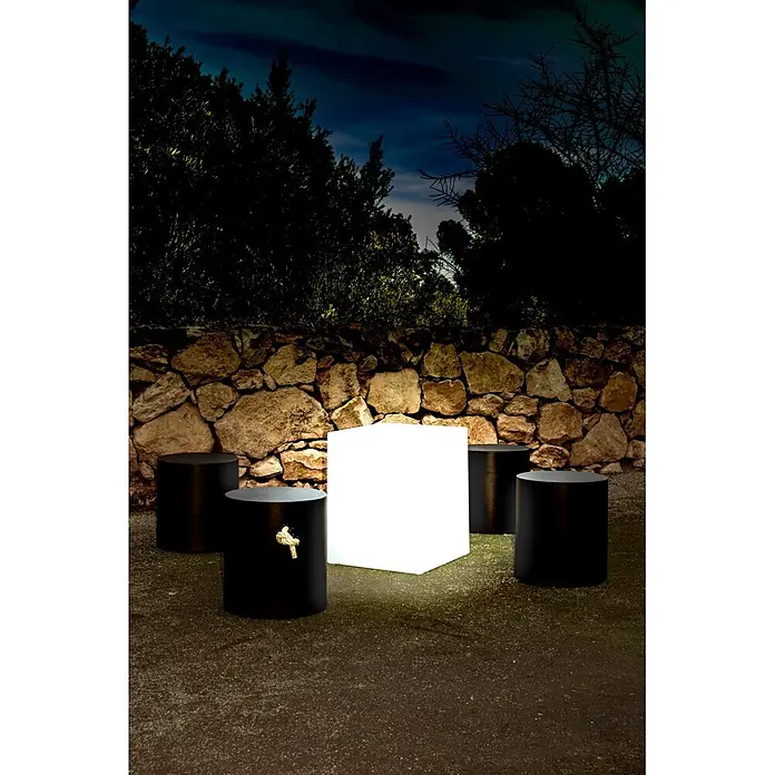 New Garden Lámpara LED decorativa Cuby 53 - Blanco, L x An x Al: 43 x 43 x 53 cm, 260 lm - Un cubo luminoso blanco y taburetes cilíndricos negros en un entorno exterior nocturno.