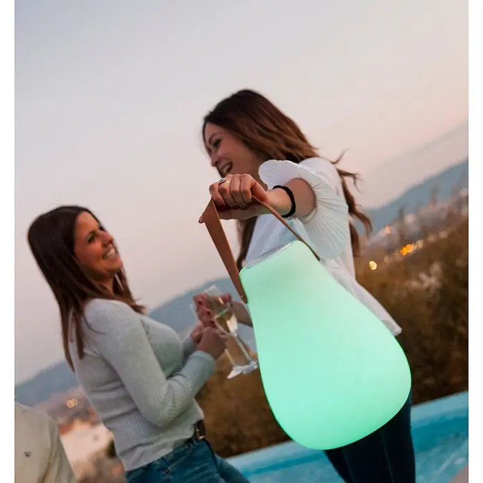 New Garden Lámpara para exterior con altavoz Kurby 38 Play - Blanco, Altavoz Bluetooth, Ø x Al: 27,5 x 37 cm, RGBW - Una mujer sostiene una lámpara portátil brillante en forma de lágrima que emite una luz verde suave, utilizada al aire libre en una reunión nocturna.