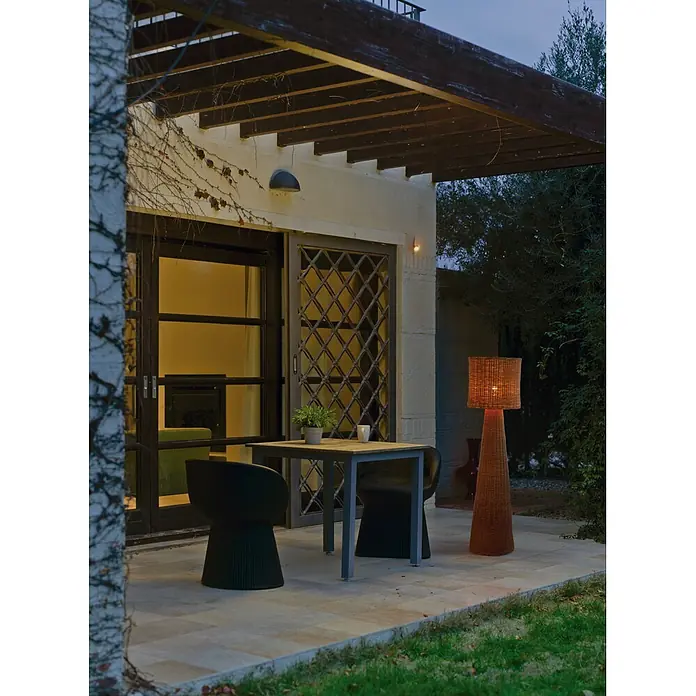 New Garden Lámpara de pie Grace 140 Nature - Altura: 137 cm - Patio exterior con lámpara de pie de ratán brillante junto a una mesa y sillas oscuras, frente a una casa y jardín al anochecer.