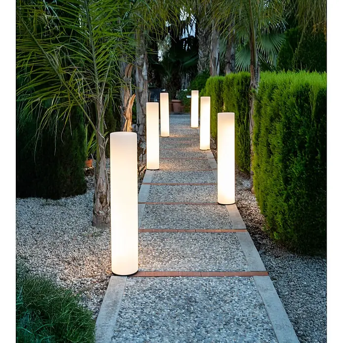 New Garden Lámpara de diseño para exterior Fity 100 - 9,5 W, Blanco, Ø x Al: 19 x 100 cm, RGB - Lámparas cilíndricas blancas iluminadas para exterior alumbran un sendero de grava entre setos y palmeras.
