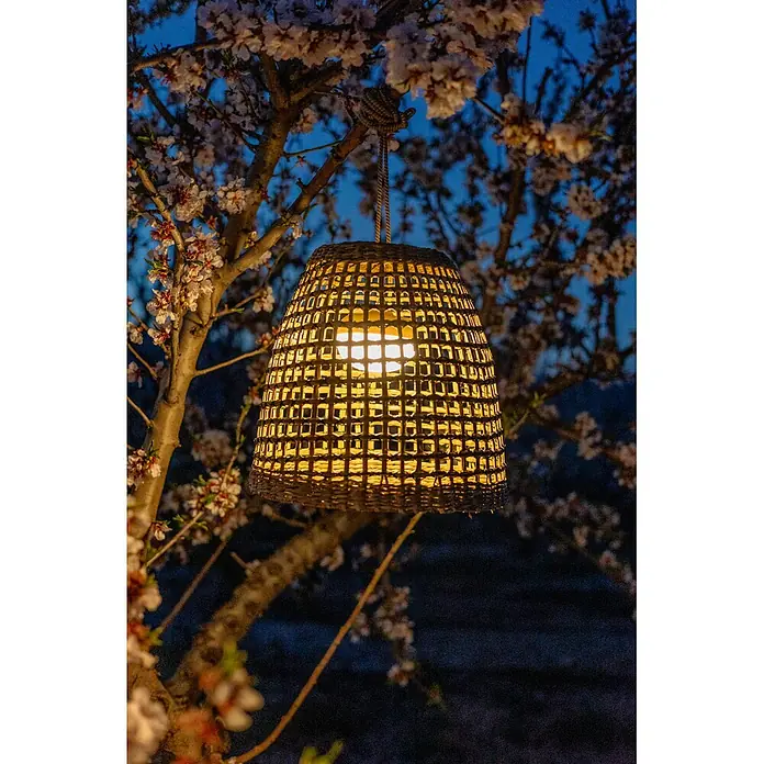 New Garden Lámpara colgante LED recargable Positano 35 - 9 W, Marrón, Ø x Al: 33 x 35 cm, Blanco cálido - Una linterna tejida brillante cuelga de un árbol florecido al anochecer, proyectando una luz cálida.