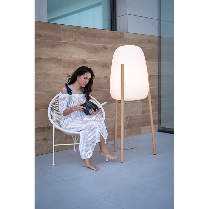 New Garden Lámpara LED decorativa Rocket 145 - 9 W, Blanco/Madera, L x An x Al: 63,5 x 63,5 x 145 cm, Blanco cálido - Una mujer lee en una silla de mimbre blanca, iluminada por una lámpara de pie grande con difusor blanco y patas de madera.