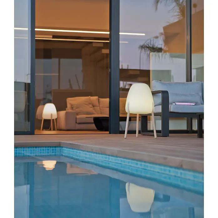 New Garden Lámpara LED decorativa Rocket 70 - 9 W, Blanco/Madera, L x An x Al: 38 x 38 x 70 cm, Blanco cálido - Una lámpara portátil blanca brillante con patas de madera sobre una terraza junto a una piscina al anochecer, reflejándose en el agua.