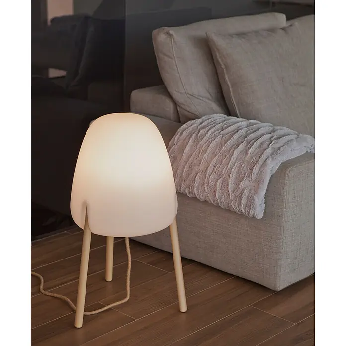 New Garden Lámpara LED decorativa Rocket 70 - 9 W, Blanco/Madera, L x An x Al: 38 x 38 x 70 cm, Blanco cálido - Lámpara de mesa blanca con patas de madera en suelo de madera, junto a un sofá gris con manta suave, ofreciendo luz ambiental.