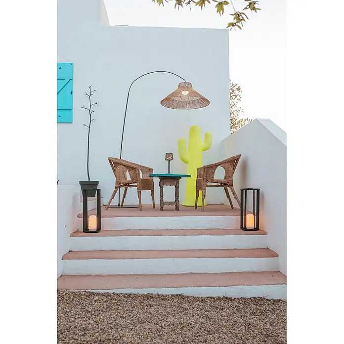 New Garden Lámpara de pie con batería Niza 230 - Marrón, 155 x 80 x 230 cm, Blanco cálido - Zona de asientos exterior con sillas de mimbre, mesa de madera, lámpara grande tejida y dos faroles negros en los escalones, junto a una escultura de cactus amarillo.