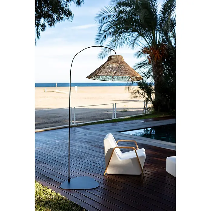 New Garden Lámpara de pie con batería Niza 230 - Marrón, 155 x 80 x 230 cm, Blanco cálido - Lámpara de pie de exterior grande con arco negro y pantalla de fibras naturales, en tarima de madera junto a silla blanca con vistas a la playa.