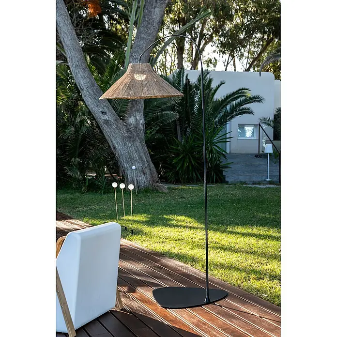 New Garden Lámpara de pie con batería Niza 230 - Marrón, 155 x 80 x 230 cm, Blanco cálido - Lámpara de pie exterior negra alta con pantalla cónica tejida, iluminando una tarima de madera.