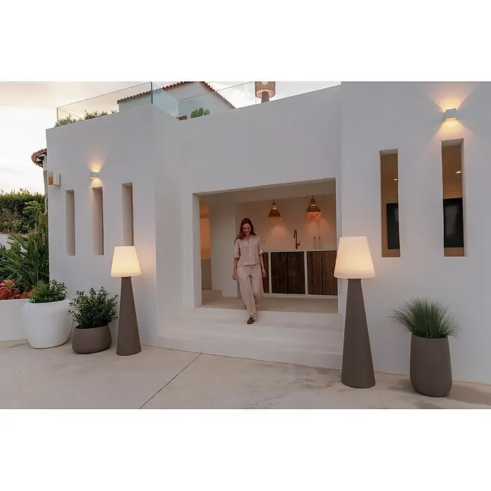 New Garden Lámpara de pie con batería Lola 165 Taupe - Taupe, Altura: 165 cm, Blanco neutro - Dos lámparas de pie exteriores altas gris oscuro iluminan una entrada de casa mientras una mujer baja las escaleras.