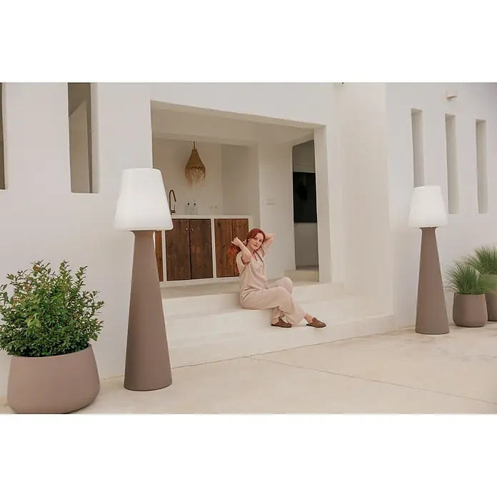 New Garden Lámpara de pie con batería Lola 165 Taupe - Taupe, Altura: 165 cm, Blanco neutro - Lámparas de pie altas de color topo con pantallas blancas y maceteros a juego con plantas verdes flanquean a una mujer sentada en escalones.