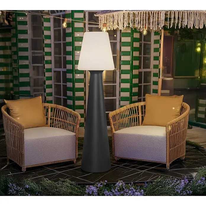 New Garden Lámpara de pie con batería Lola 165 Antracita - Antracita, Altura: 165 cm, Blanco neutro - Una lámpara de pie cónica alta, gris oscuro y con pantalla blanca, ilumina un patio entre dos sillas de mimbre.
