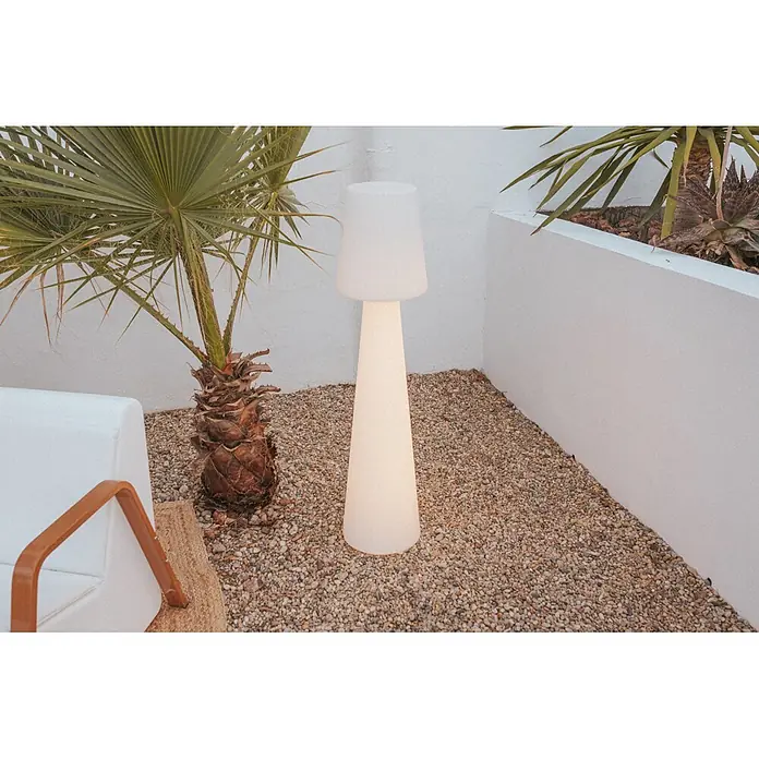 New Garden Lámpara de pie con batería Lola 110 - Blanco, Altura: 110 cm, RGBW - Una lámpara de pie alta, blanca e iluminada para exteriores, sobre grava junto a una palmera.