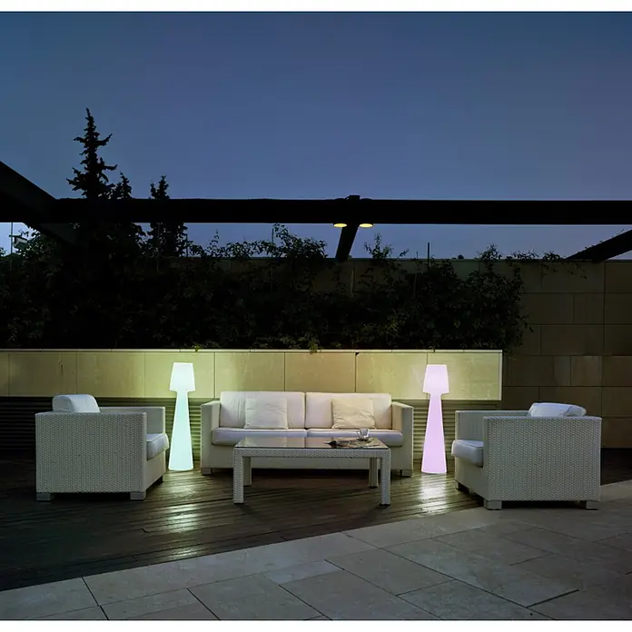 New Garden Lámpara de pie con batería Lola 110 - Blanco, Altura: 110 cm, RGBW - Dos lámparas de pie altas e iluminadas, una azul verdosa y una rosa, iluminan un patio exterior con muebles blancos.