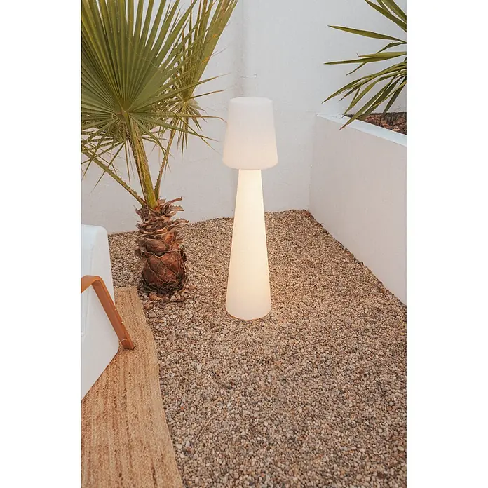 New Garden Lámpara de pie con batería Lola 110 - Blanco, Altura: 110 cm, RGBW - Una lámpara de pie exterior alta y blanca luminosa en un patio de grava junto a una palmera.